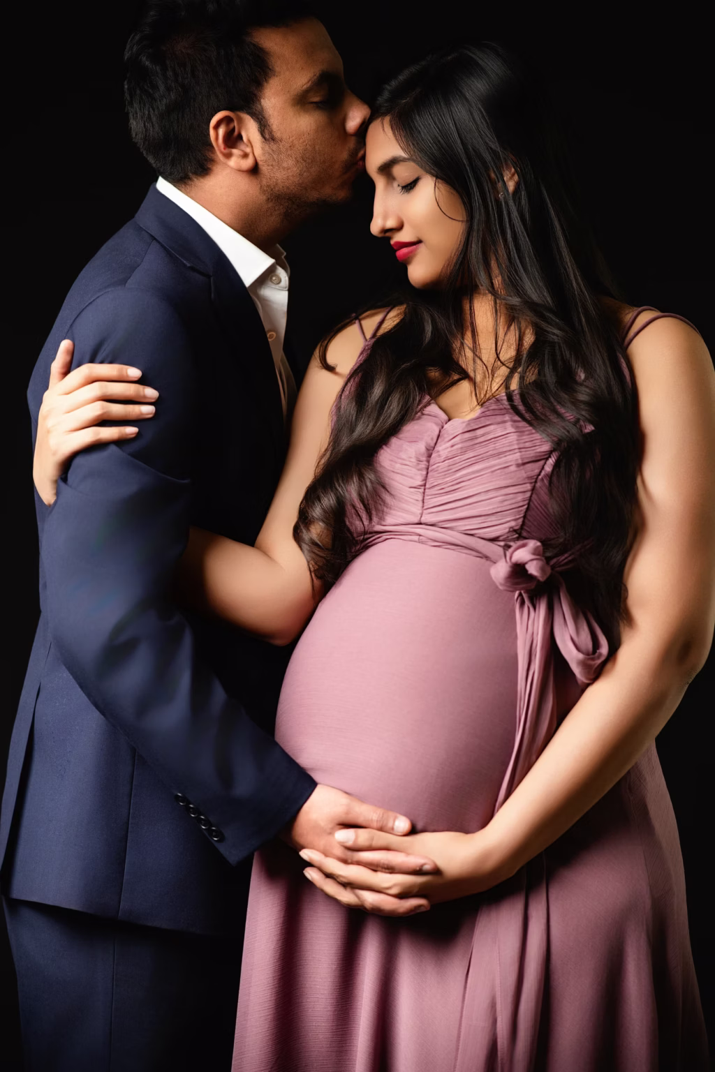 Close-up studio portrait against black background, man in navy suit kissing forehead of woman in mauve-colored maternity dress, both with eyes closed creating peaceful tender moment - couple pregnancy photos, forehead kiss intimate