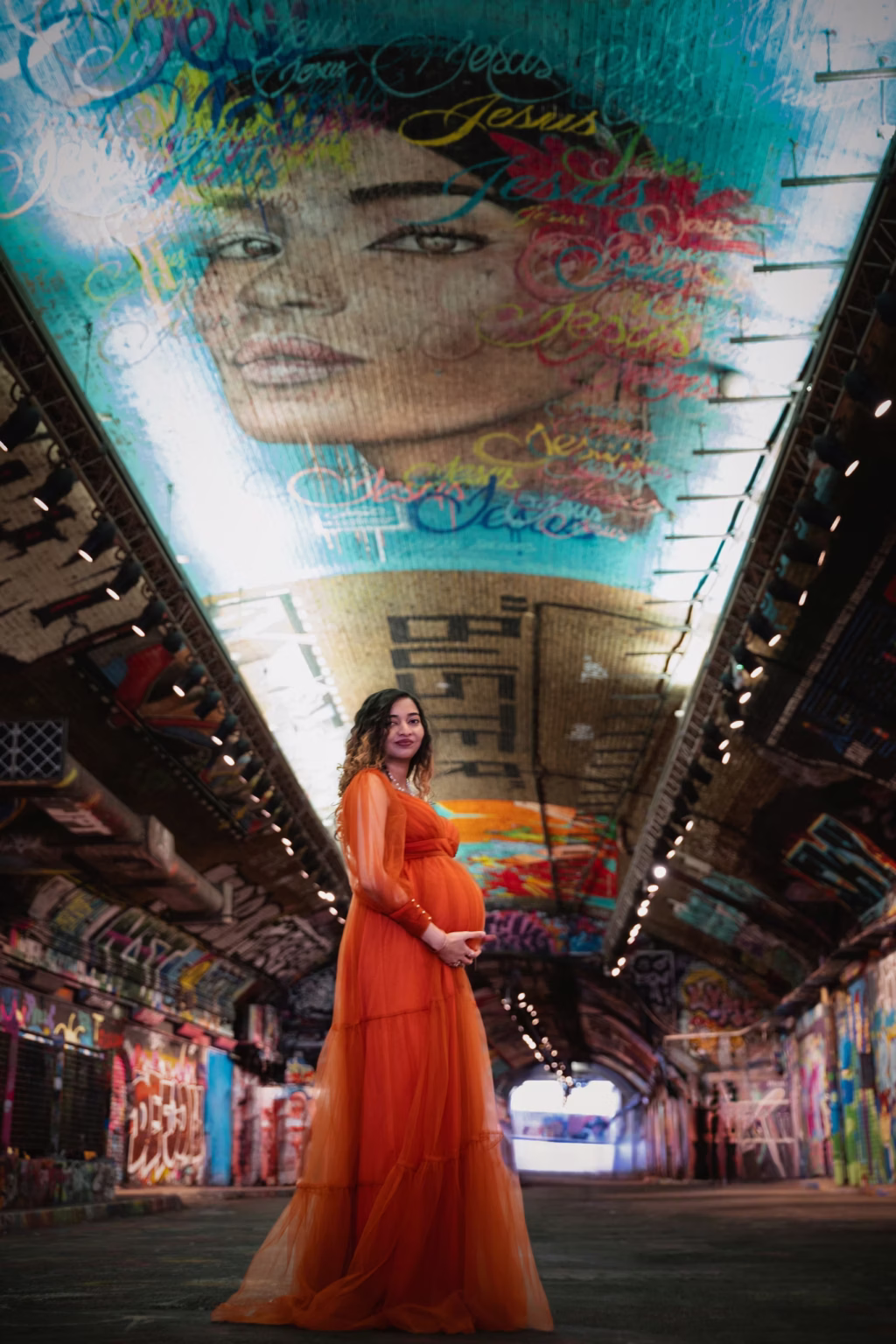 Vibrant shot inside Leake Street Graffiti Tunnel, woman in orange tulle gown standing in center of tunnel, large colorful graffiti mural of woman's face looking down from curved ceiling - maternity photoshoot package, graffiti tunnel art street
