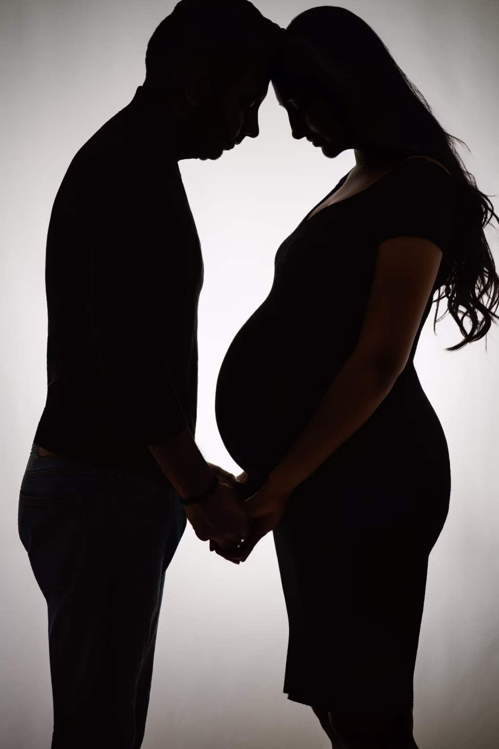 Stark artistic silhouette of couple facing each other against bright white light, foreheads touching, holding hands low between them, highlighting sharp outline of woman's pregnant profile - london maternity photoshoot silhouette intimacy