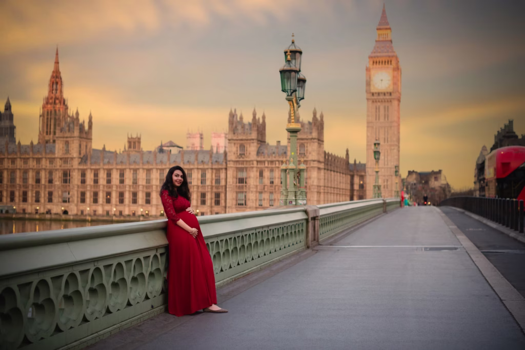Expansive shot on Westminster Bridge, woman in long red gown leaning against ornate bridge railing, Houses of Parliament and Big Ben stretching across background under dramatic cloudy sunset - photographers maternity, Westminster Bridge sunset