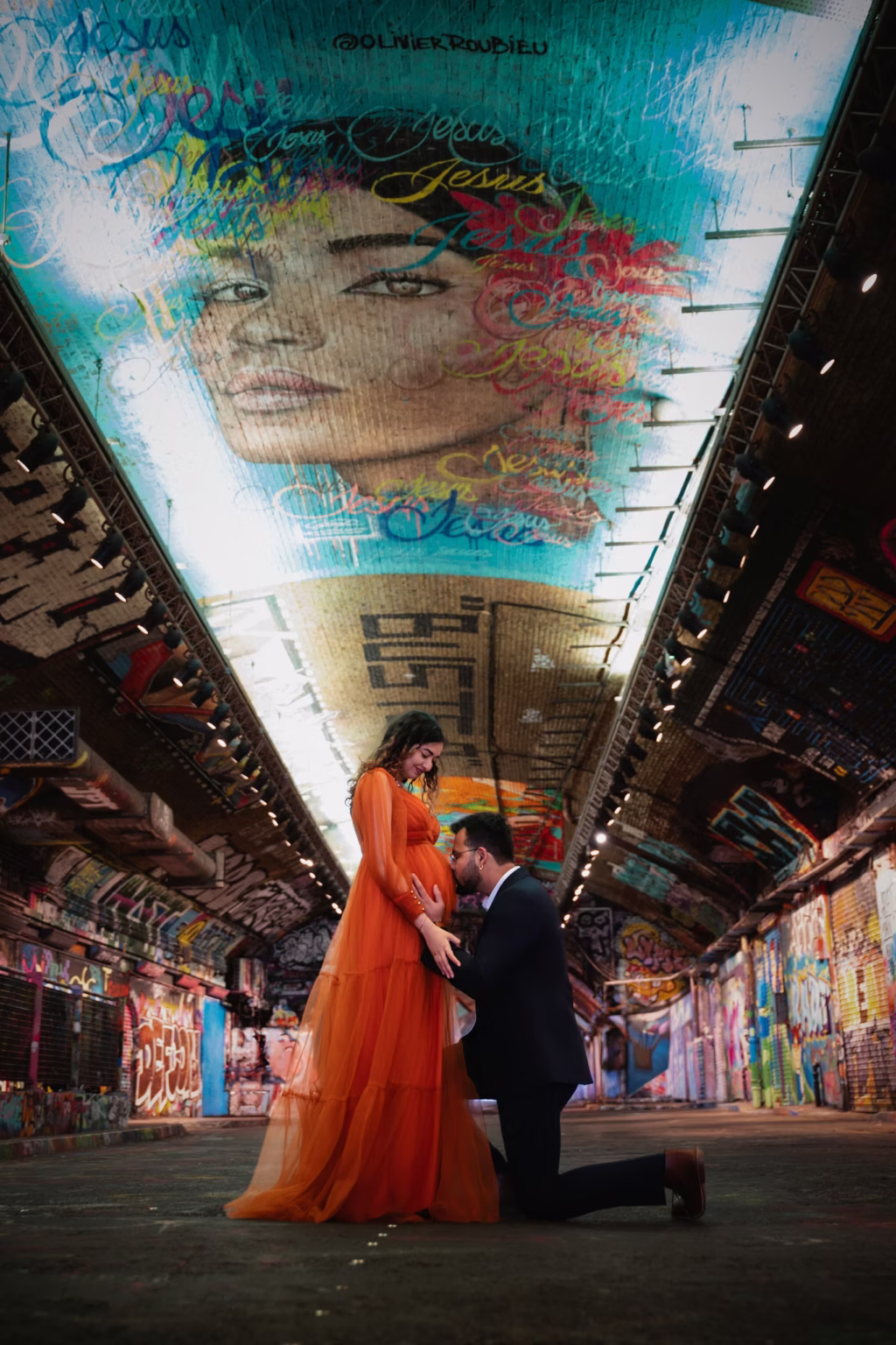 Urban artistic shot in Leake Street Tunnel, woman in vibrant orange gown standing, man kneeling before her kissing her belly, tunnel ceiling covered in elaborate blue and red graffiti art - maternity photographer, Leake Street graffiti belly kiss urban