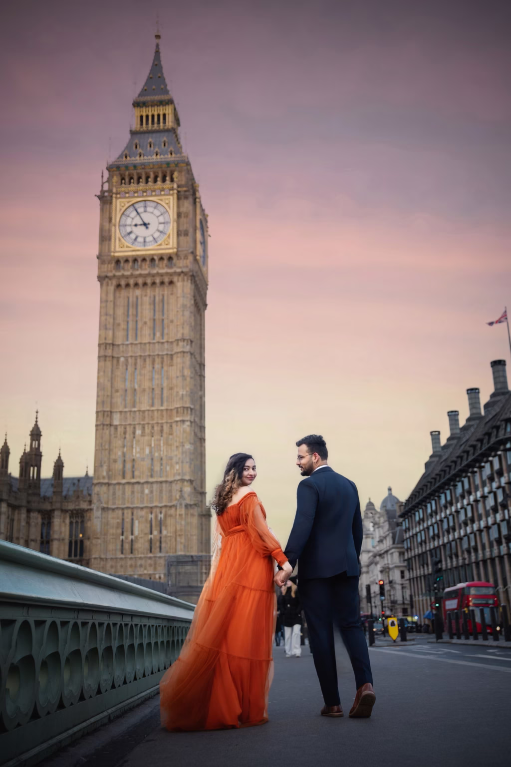 Cinematic shot on Westminster Bridge at twilight, couple walking away from camera toward Big Ben holding hands, woman in flowing orange tulle gown looking back over shoulder with smile - photographer maternity, Westminster Bridge walking away