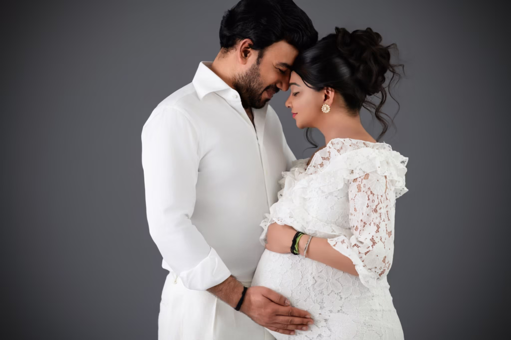 Soft studio portrait against dark grey background, man in white button-down shirt and woman in delicate white lace maternity dress with eyes closed and foreheads touching - maternity studio photoshoot, photoshootshoot white lace soft intimate