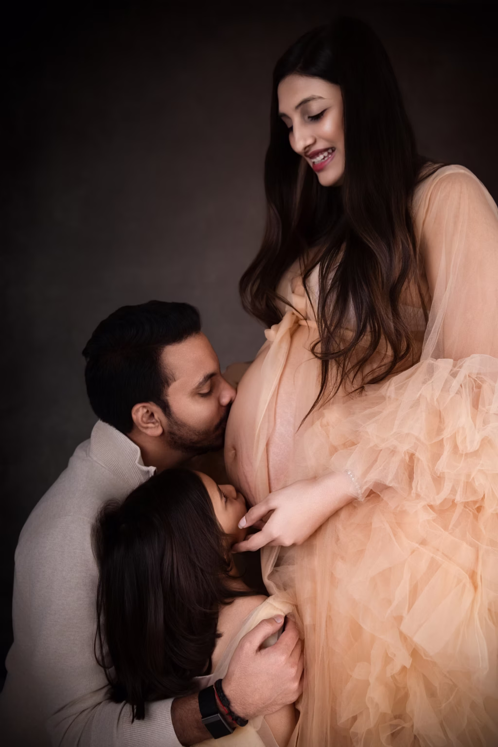 Warm intimate family portrait against dark textured background, woman in ruffled peach-colored tulle gown, man and young girl leaning in to gently kiss her bare pregnant belly - maternity studio photoshoot, affection kiss belly