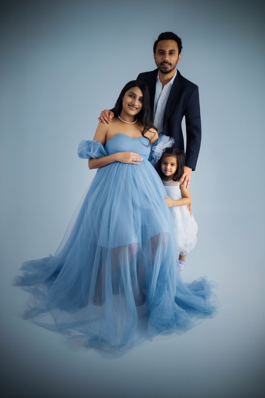 Family portrait in studio against light blue background, mother seated in voluminous light blue tulle gown, father stands behind her in suit, young daughter in white dress stands beside mother leaning against tulle gown - pregnancy shoot studio, photoshootshoot family coordinated styling