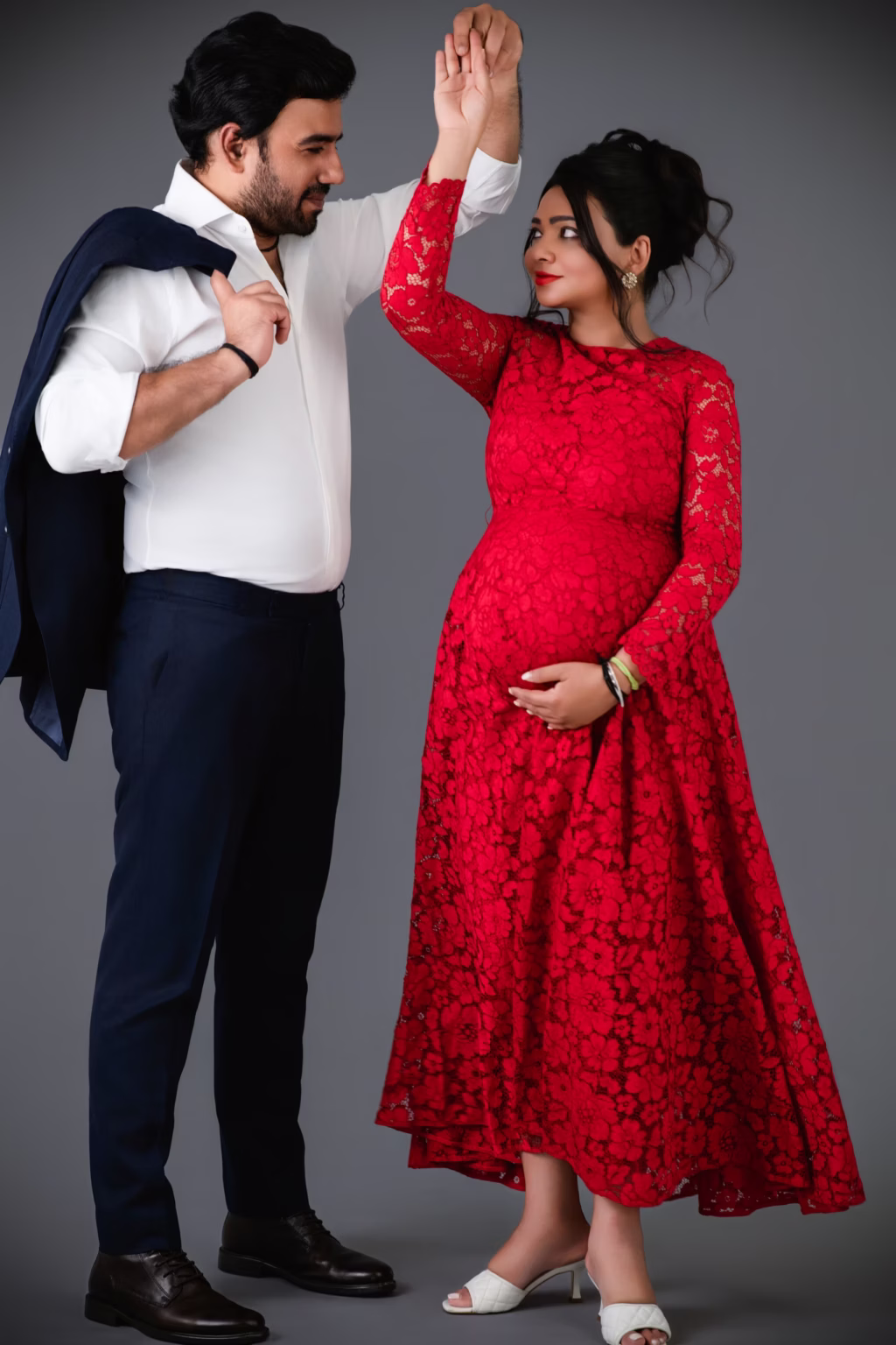 Full-length studio shot against grey background, pregnant woman in red lace gown "twirling" as partner holds her hand upward, man dressed in white shirt and navy trousers carrying suit jacket over shoulder - maternity shoot studio, red lace gown twirl