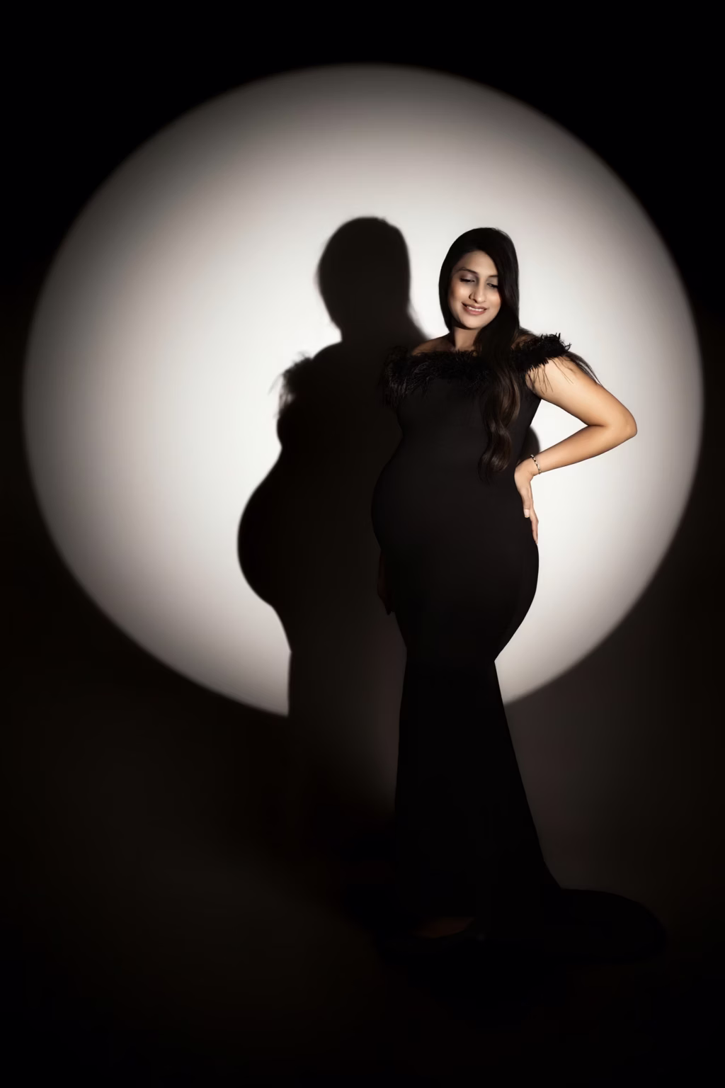 Dramatic studio portrait using circular "spotlight" effect on black background, pregnant woman standing in profile wearing sleek black gown with feathered neckline, silhouette sharply defined against light with hand on hip - studio maternity photography, black feathered spotlight profile