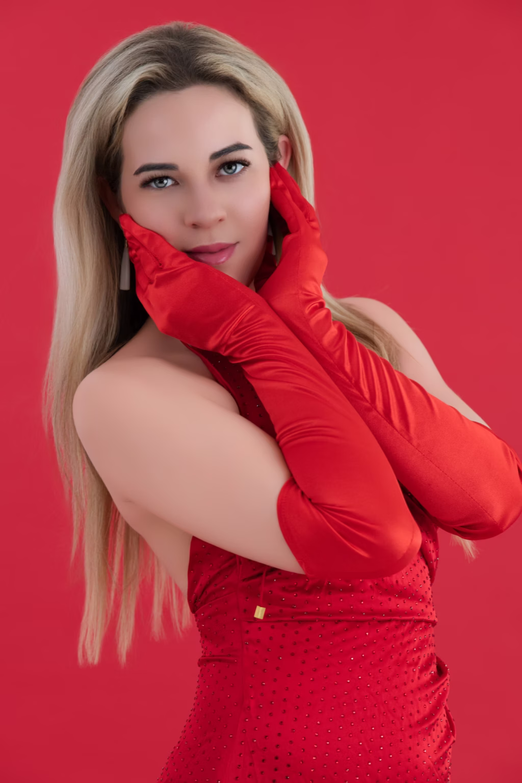 London model poses - fashion photoshoot, blonde woman red background red studded dress long red satin gloves holding gloved hands framing face light-colored eyes neutral makeup elegant