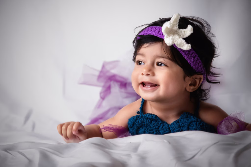Bright studio portrait of smiling infant girl in purple crocheted headband with large white starfish accent and teal crocheted mermaid top, lying on soft white fabric background - london new born photoshoot mermaid starfish smile