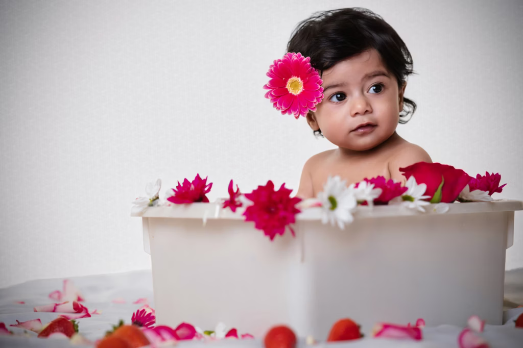 Creative milk bath style portrait, infant girl sitting in white tub filled with white liquid topped with vibrant pink and white flowers, large pink flower behind ear, strawberries scattered on white surface - newborn photography shoot, milk bath flowers