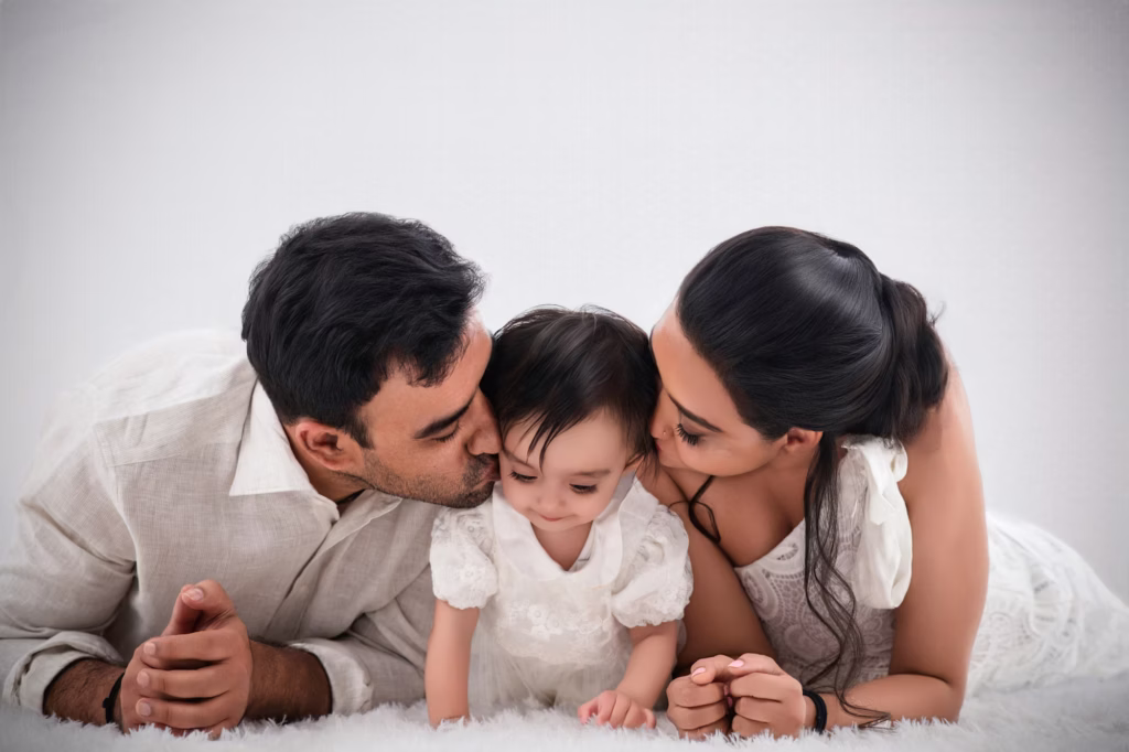 Tender family portrait against white background, father and mother lying on white shag rug both leaning in to kiss young daughter on cheeks, daughter wearing white lace dress looking down - london newborn photography family kiss