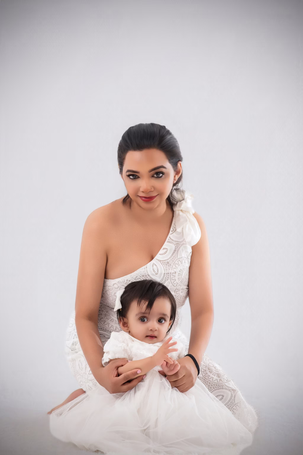 High-key studio portrait of mother and infant daughter both dressed in white, mother in one-shoulder lace dress, baby in ruffled tutu dress with white bow, both looking directly at camera - newborn photos, mother daughter white lace