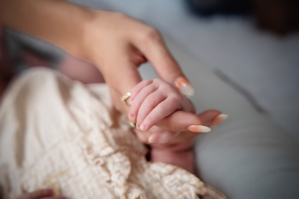 Tender close-up shot of newborn's tiny hand grasping adult's finger, adult wearing decorative gold ring and manicured nails, softly blurred background emphasizing delicate scale - newborn photo sessions, hand grasp gold ring