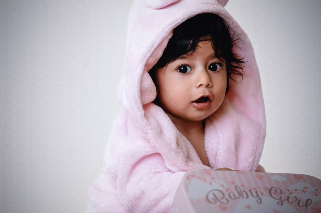 Cute studio portrait of baby girl in plush pink hooded robe with animal ears, wide dark eyes and curious expression, looking over edge of pink box saying Baby Girl - london newborn photography pink robe animal ears box