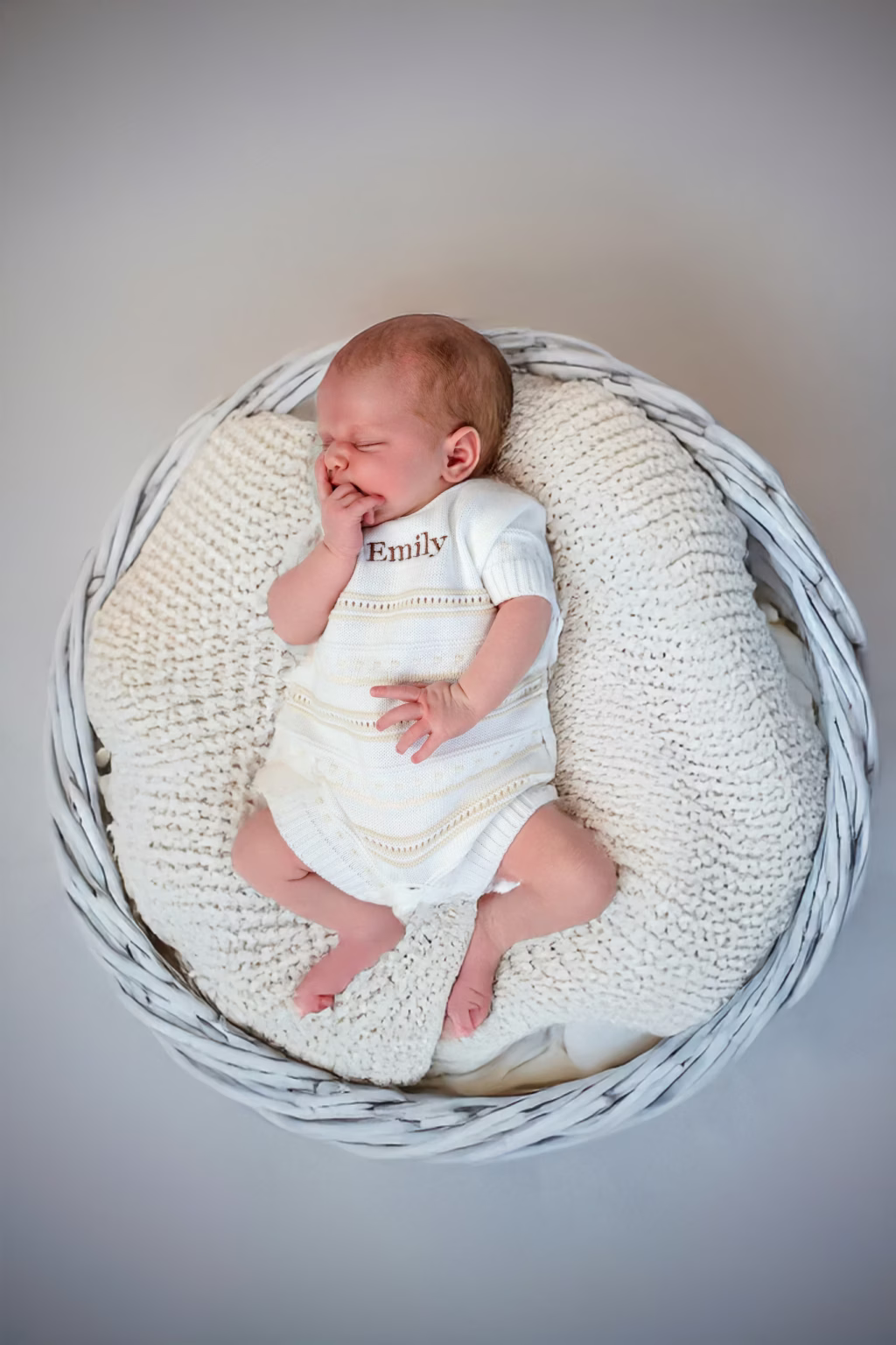 Serene newborn portrait of baby Emily sleeping soundly in white wicker basket, lying on thick cream-colored knit blanket wearing personalized white knit romper with name embroidered on chest - new born photos, emily basket personalized