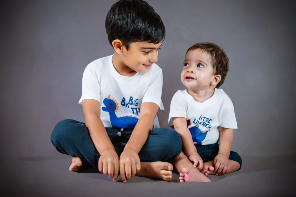 Version of two brothers from previous image sitting on grey background wearing matching Big Brother and Little Brother dinosaur t-shirts older boy looking affectionately at younger sibling - family studio photoshoot