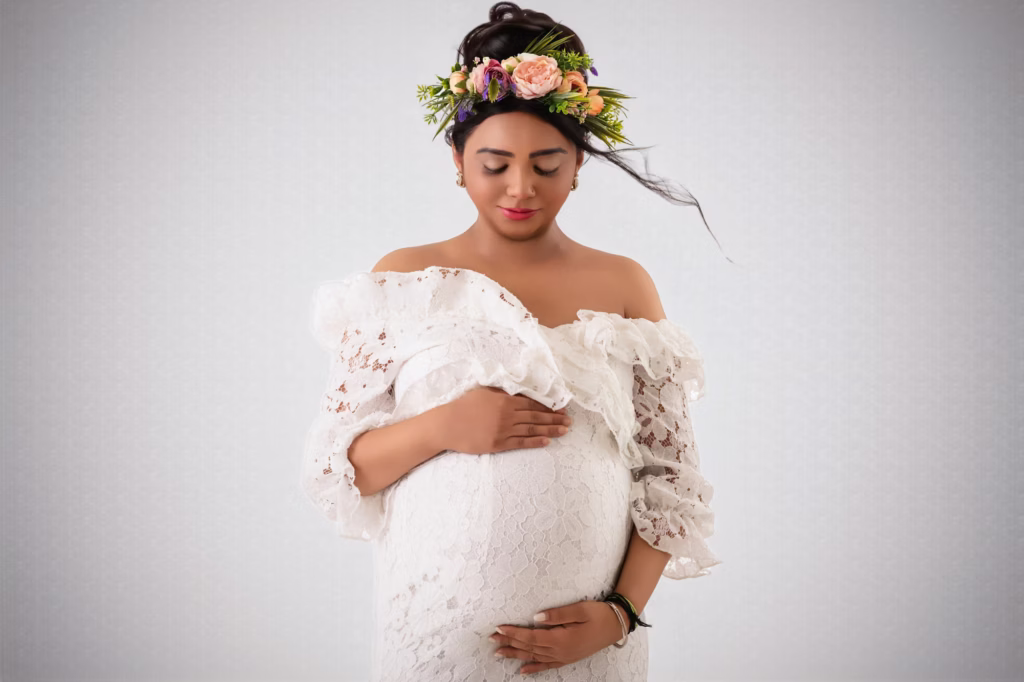 Ethereal studio portrait of woman in white lace off-the-shoulder dress, wearing lush flower crown made of pink and orange roses, eyes closed looking down toward bump, hair caught in light breeze - pregnancy shoot studio, for maternity ethereal white lace rose crown breeze
