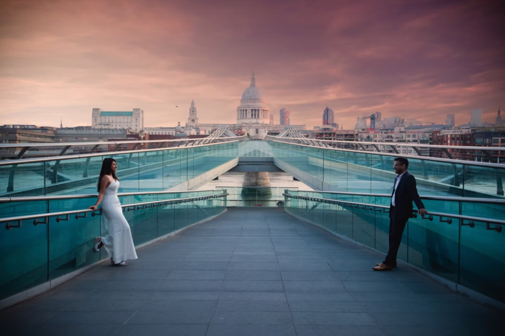 For engagement very wide symmetrical shot of Millennium Bridge with couple separated on far left and right sides each leaning against opposite railings looking toward one another with St. Paul's Cathedral perfectly centered in distance - best engagement photos