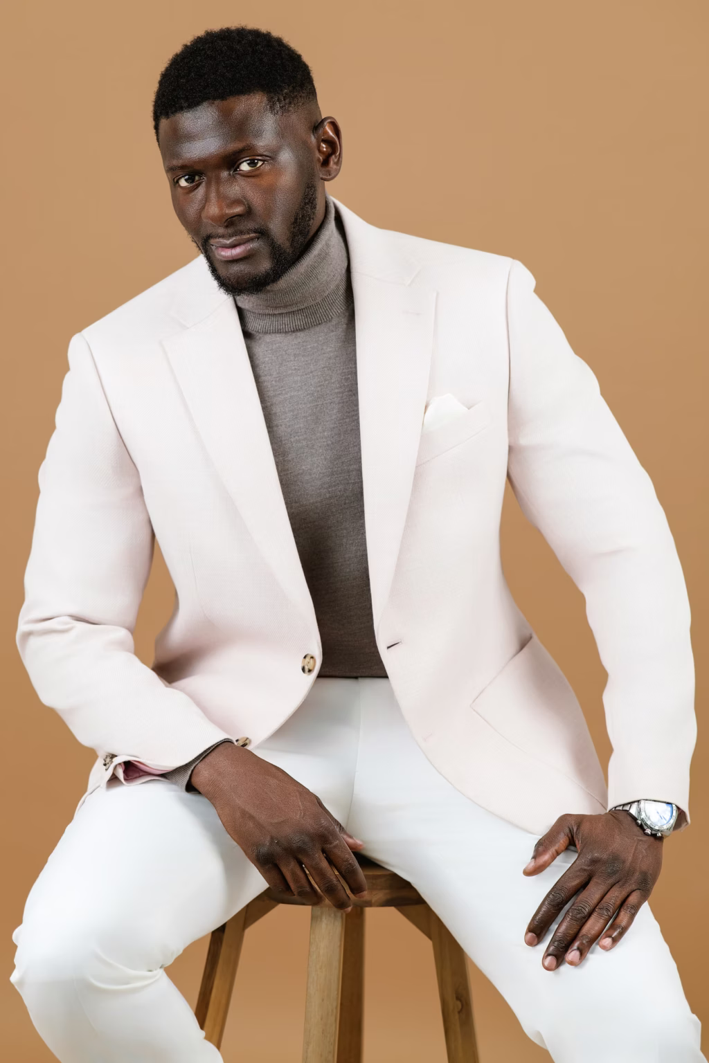 Sophisticated seated London Photography And Fashion portrait of man wearing light pink blazer and taupe turtleneck against neutral tan background