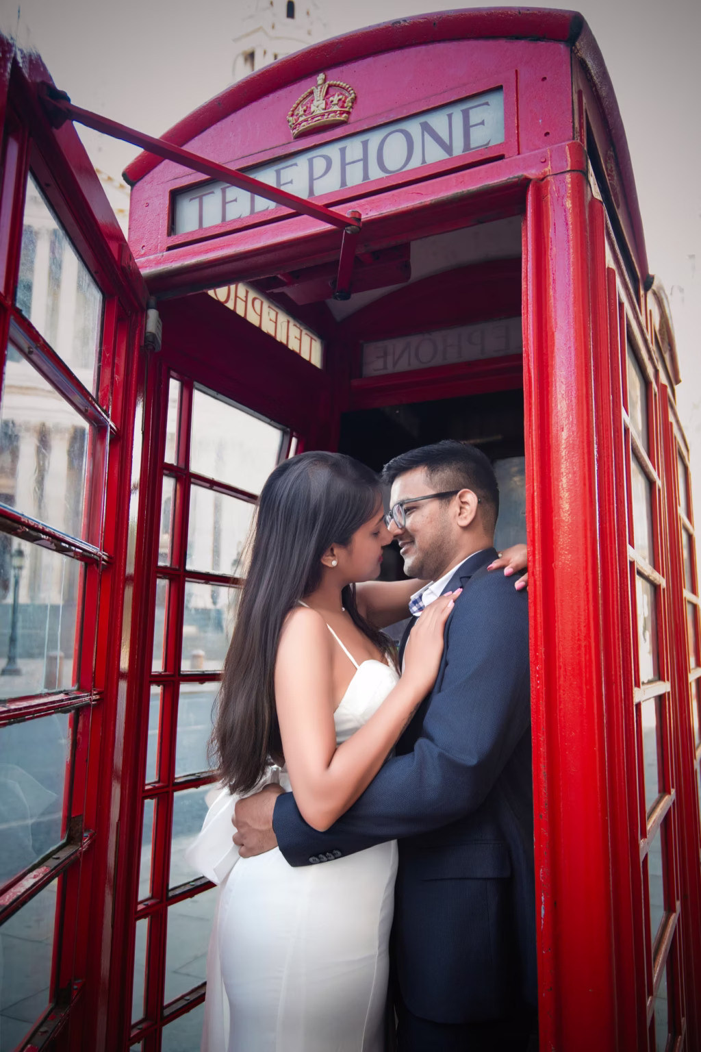 Pre wedding photoshoot intimate portrait of couple inside iconic red London telephone box, man in navy suit and glasses with woman in white spaghetti-strap gown standing close together almost touching noses - pre wedding photoshoot london