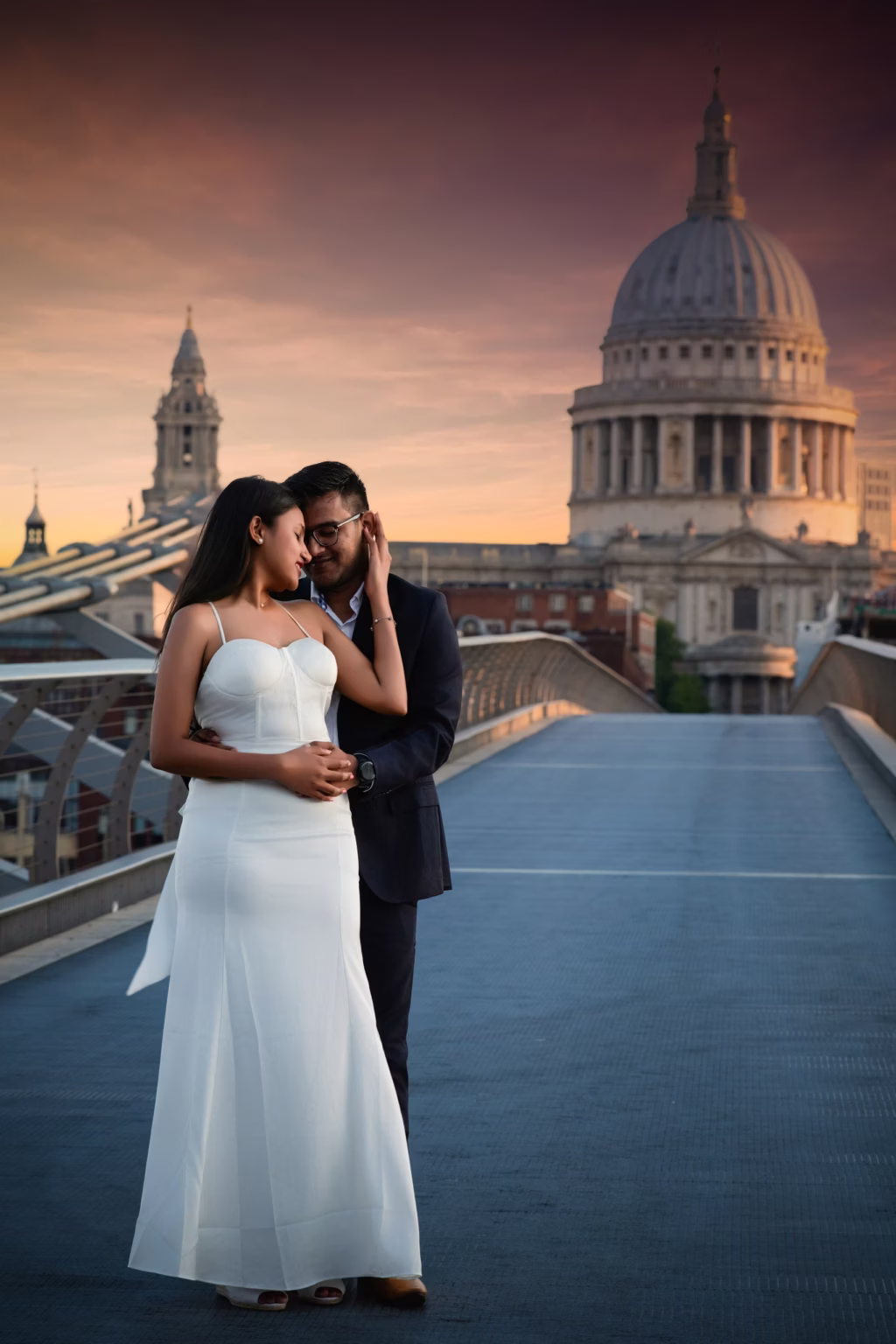Pre wedding photoshoot romantic wide shot of couple on Millennium Bridge with man standing behind woman with arms wrapped around her waist, woman reaching back to touch his face against majestic St. Paul's Cathedral dome - proposal photographer london