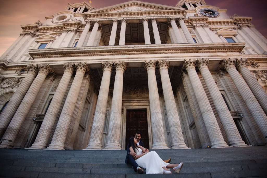 Pre wedding shoot cinematic low-angle shot of couple sitting on wide stone steps of St. Paul's Cathedral with massive Corinthian columns and grand cathedral facade looming overhead, rich warm twilight sky - engagement shoot