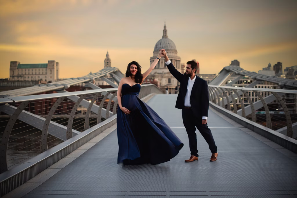 Dramatic outdoor shot on Millennium Bridge, pregnant woman in flowing strapless navy blue gown "dancing" with partner in navy suit, St. Paul's Cathedral centered in background under golden sunset sky - maternity photoshoot cost, Millennium Bridge dance Cathedral