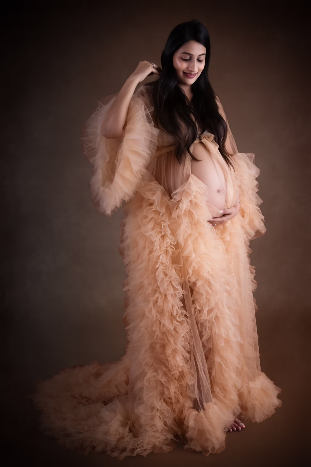 Full-length studio portrait of pregnant woman wearing dramatic floor-length peach tulle robe with heavy ruffling and long train, looking down at her exposed belly through sheer front garment against dark brown backdrop - photographer maternity, peach robe dramatic