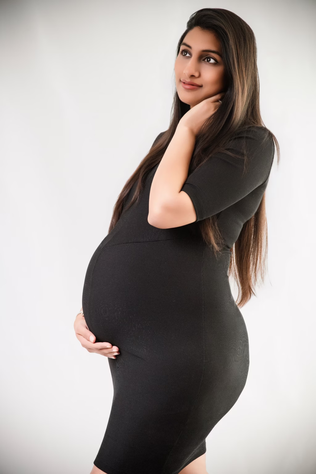 Studio portrait of pregnant woman in form-fitting textured black dress against light grey background, standing in profile to showcase bump, one hand resting on neck other on belly, looking off-camera with thoughtful expression - maternity photographer, profile textured dress