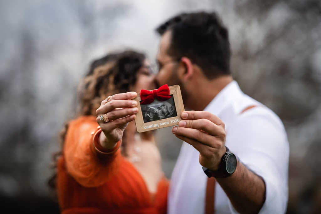Creative announcement photo, two hands in foreground holding small cardboard frame containing ultrasound photo with red bow and text "coming soon 2026," couple visible but blurred in background sharing a kiss - london pregnancy photography ultrasound announcement coming soon