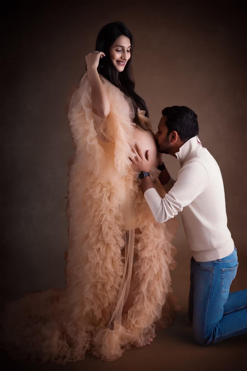 Studio portrait against dark brown backdrop, pregnant woman wearing voluminous peach ruffled gown, partner kneeling on one knee in cream sweater and jeans affectionately kissing her exposed baby bump - maternity studio photoshoot, peach gown kneeling kiss