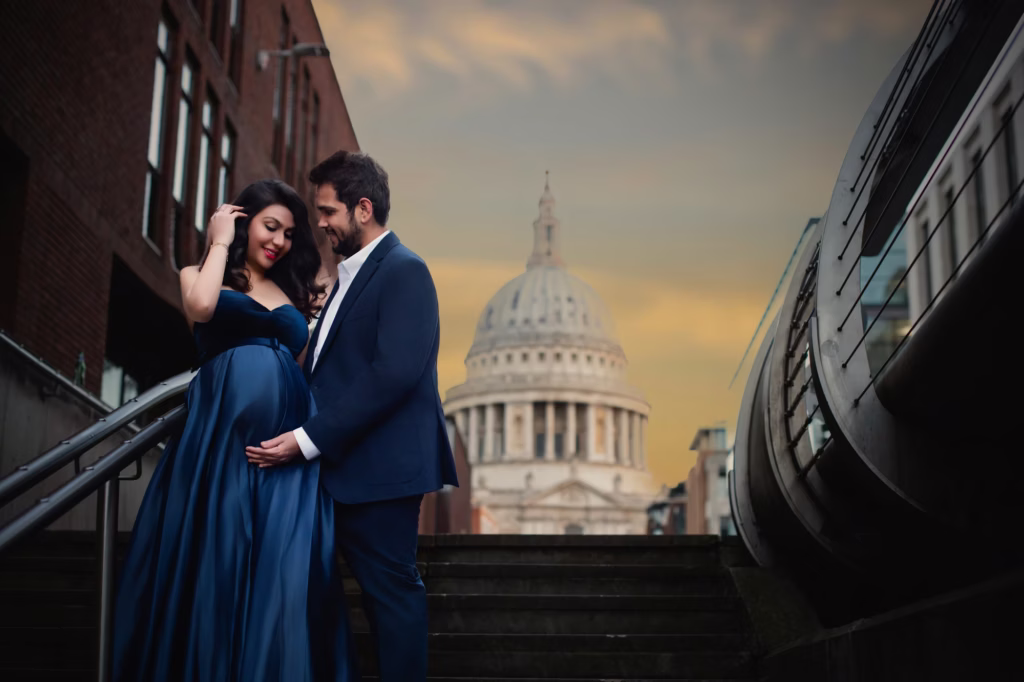 Dramatic outdoor shot at dusk, couple standing on stone stairs with brick building to left, woman in flowing navy gown, man in dark suit, St. Paul's Cathedral perfectly framed in background under golden sky - couple pregnancy photography, stone stairs St Paul's Cathedral dusk