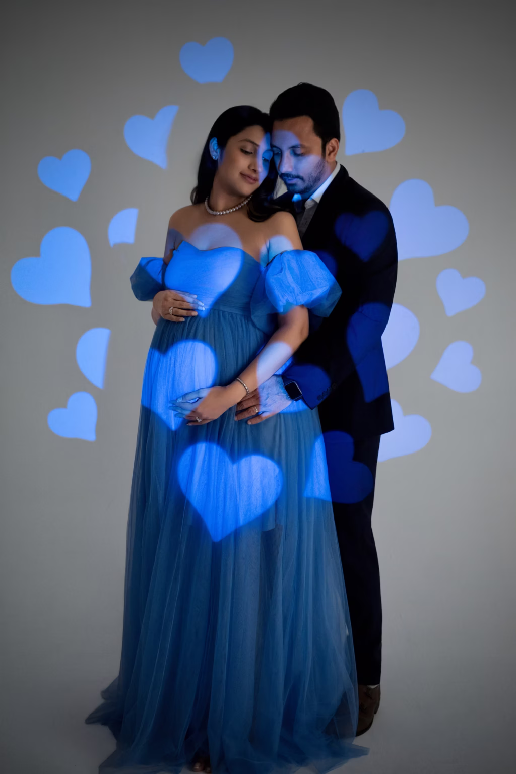 Artistic studio shot featuring couple in formal wear, woman in blue tulle gown, man in suit, standing close together with eyes closed, blue heart-shaped light patterns projected across them and background - maternity photographer london, blue tulle hearts projected light