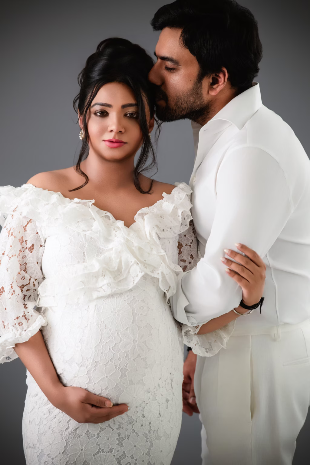 Close-up studio portrait against grey backdrop, woman in white lace dress with ruffled neckline looking directly at camera with serious expression, man in white dress shirt standing behind gently kissing top of her head - couple pregnancy photoshoot, white lace serious kiss head