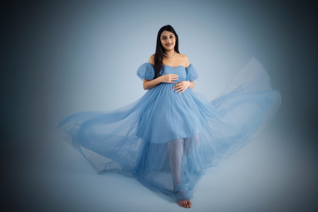 High-fashion studio shot against light blue backdrop, woman wearing light blue tulle gown with off-the-shoulder puff sleeves, sheer fabric of skirt caught in fan or tossed creating flowing ethereal "wind" effect - pregnancy photoshoot cost, light blue tulle flowing