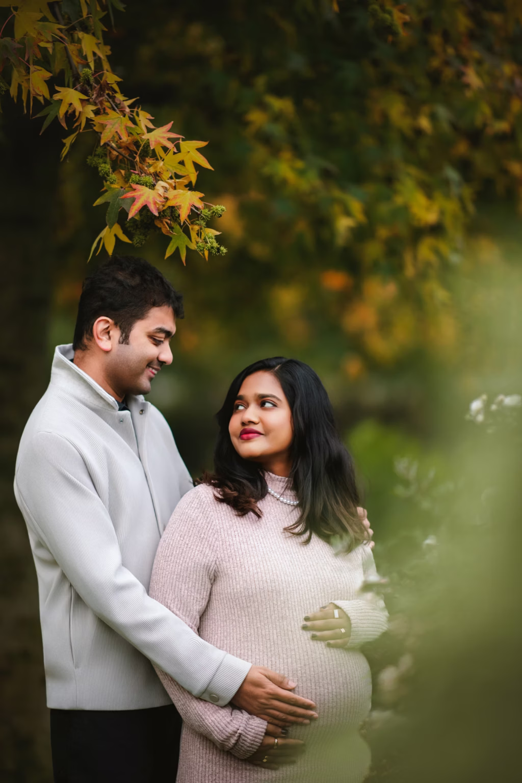 Warm outdoor portrait of expectant couple with hands on baby bump framed by autumn leaves, man in light grey ribbed jacket, woman in light pink sweater and pearl necklace - london pregnancy shoot autumn couple hands bump