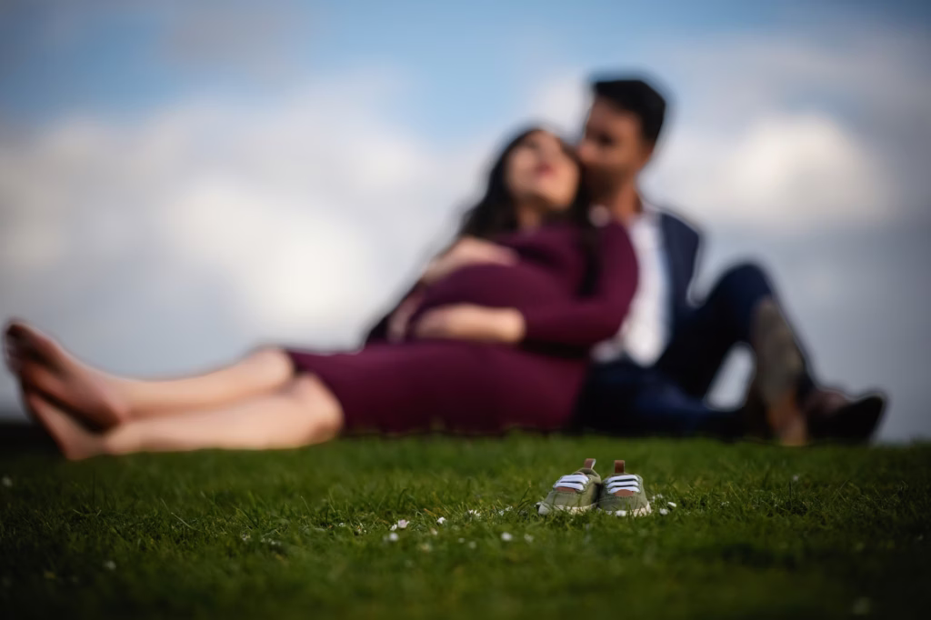 Artistic pregnancy portrait with tiny green baby shoes in sharp foreground and expecting couple blurred in park background - london pregnancy shoot baby shoes depth field