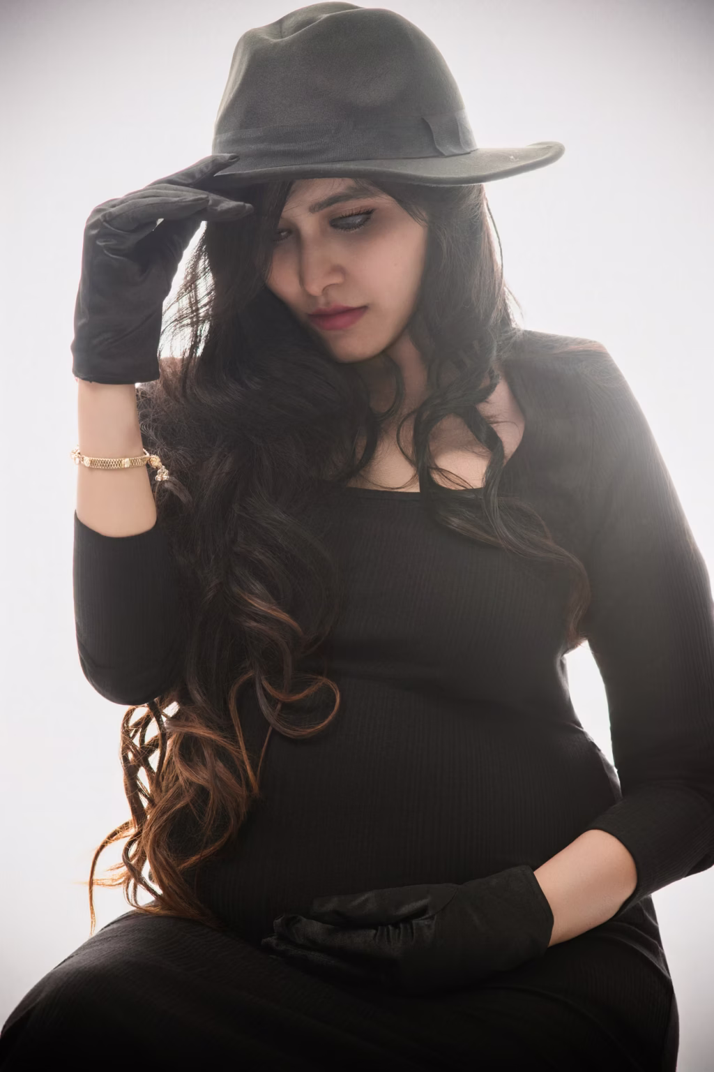 High-fashion studio portrait of expectant mother in black long-sleeved dress, black gloves and black fedora, looking down pensively adjusting hat brim - london pregnancy shoot fashion black fedora