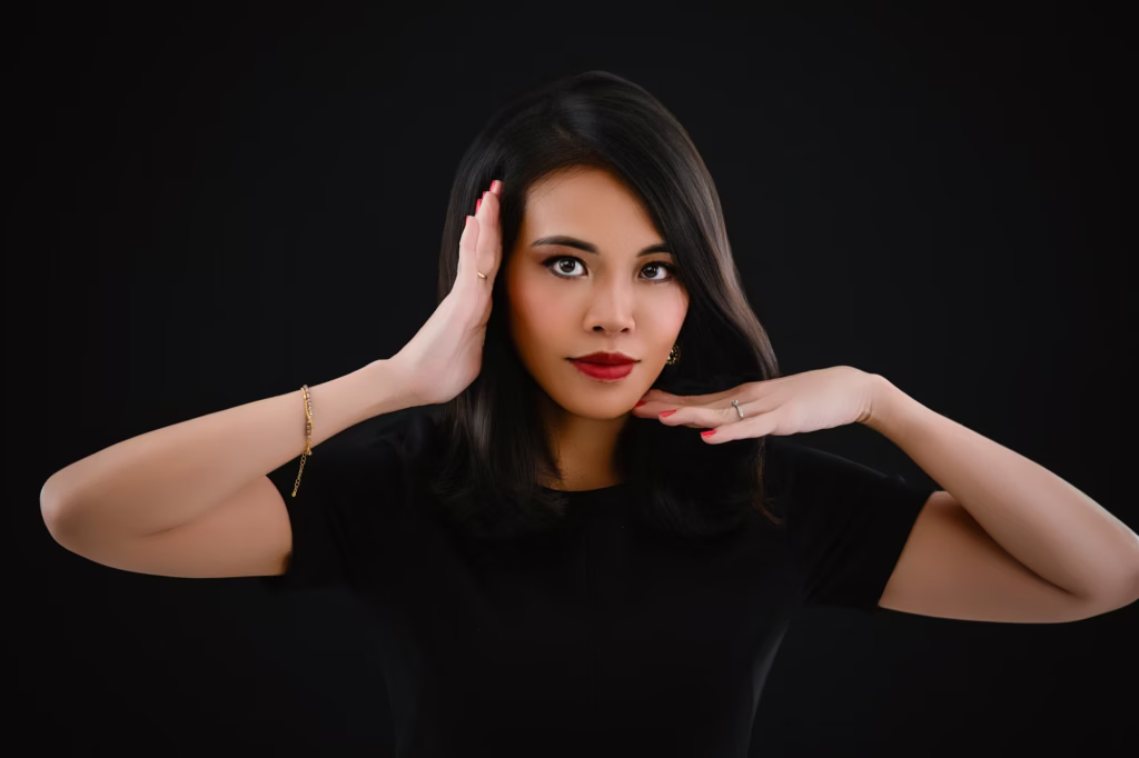 London Fashion Photographers - Centered studio portrait woman black background black crewneck top hands raised face symmetrical elegant pose bold red lip gold jewelry bracelet ring