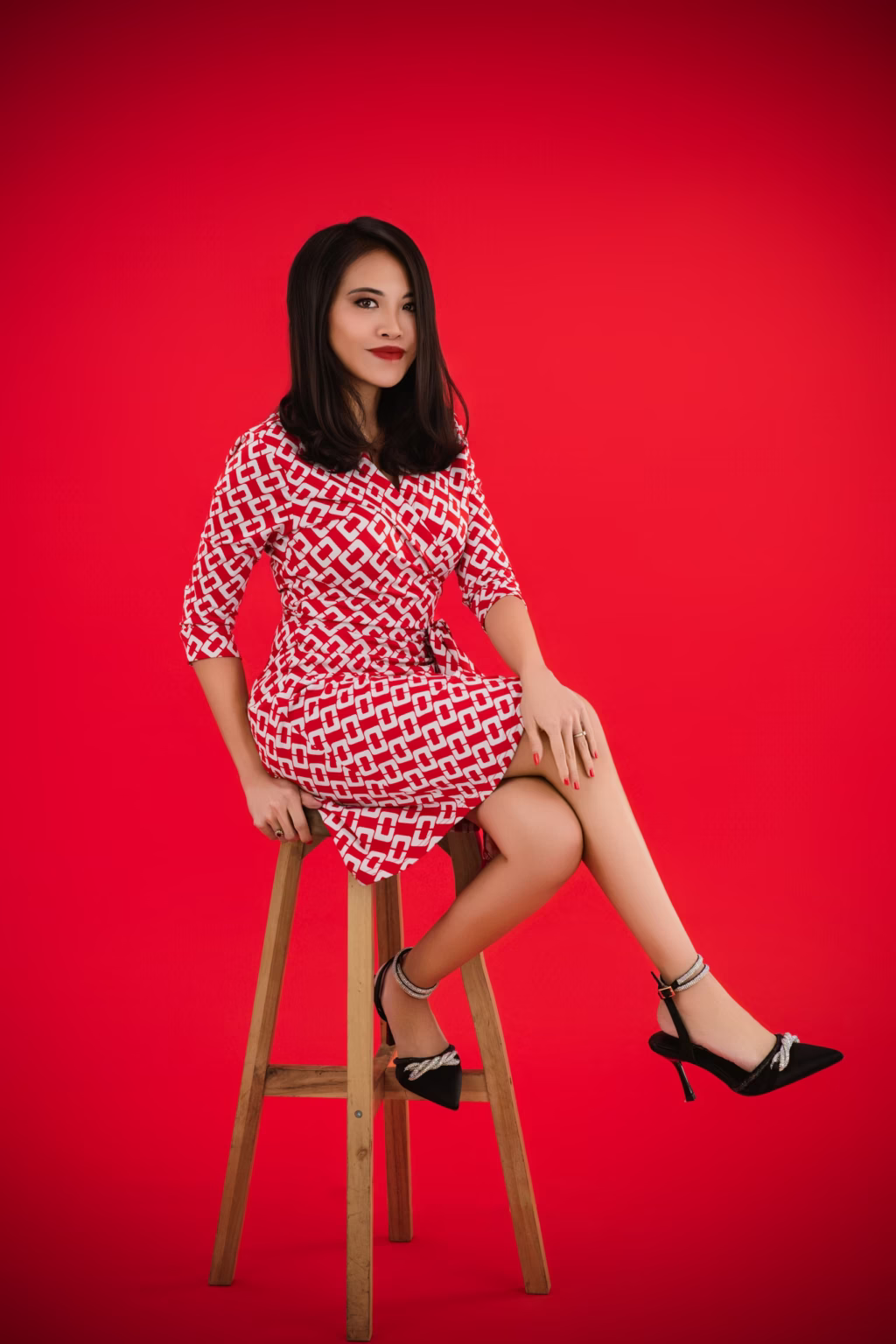 London professional model photoshoot - fashion photographer, Full-length studio shot woman sitting high wooden stool vibrant red background red white geometric print wrap dress black high-heeled sandals silver embellishments