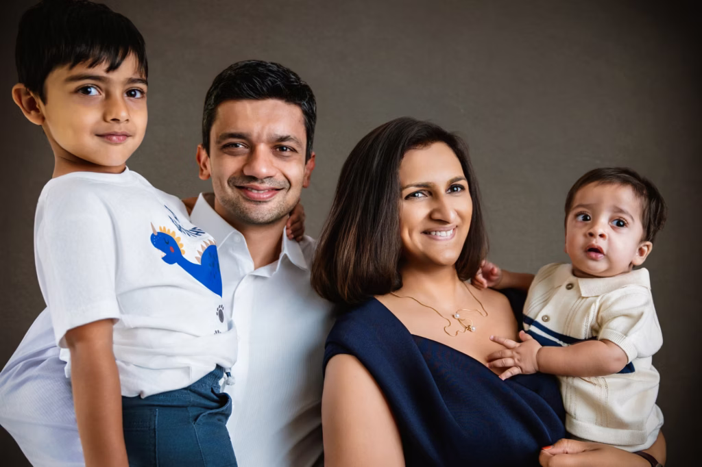 Formal studio portrait of family of four against dark grey background parents and older son smiling while mother holds baby dressed in white and navy - family portrait studio