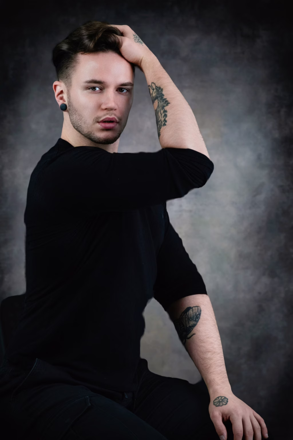 Moody studio portrait of man in black long-sleeved shirt running hand through hair with direct gaze, showcasing woman's face tattoo on inner forearm and hand flower tattoo - fashion photoshoot