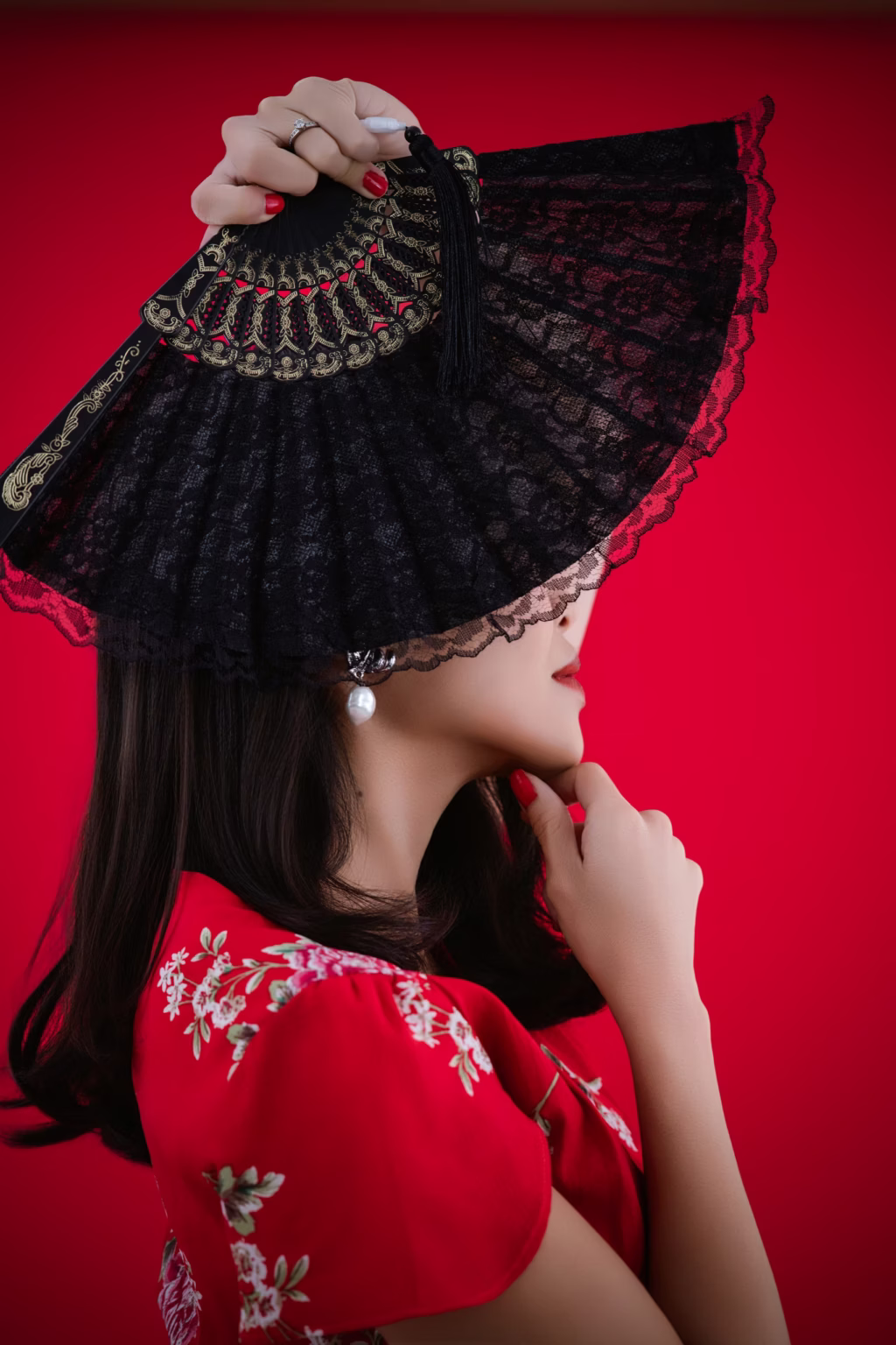 London Fashion Photography London striking profile-view portrait of woman against bold red background holding large black lace folding fan, traditional-style red dress with white floral embroidery and pearl earring