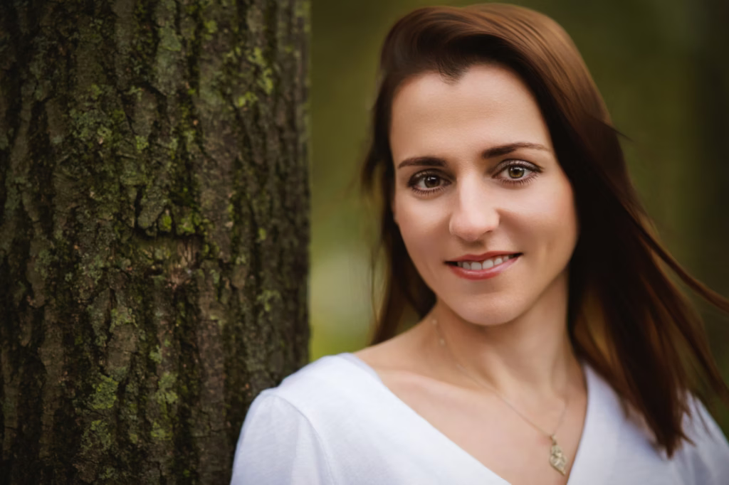 Close-up lifestyle portrait of woman with brown hair leaning against textured tree bark in white V-neck top with gentle natural expression - model shoot