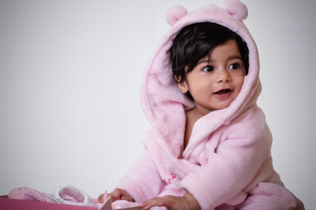 Bright studio portrait of infant girl in plush pink hooded robe with animal ears, dark hair and large expressive brown eyes, happy open-mouthed expression looking off-camera - newborn photographer london pink robe happy
