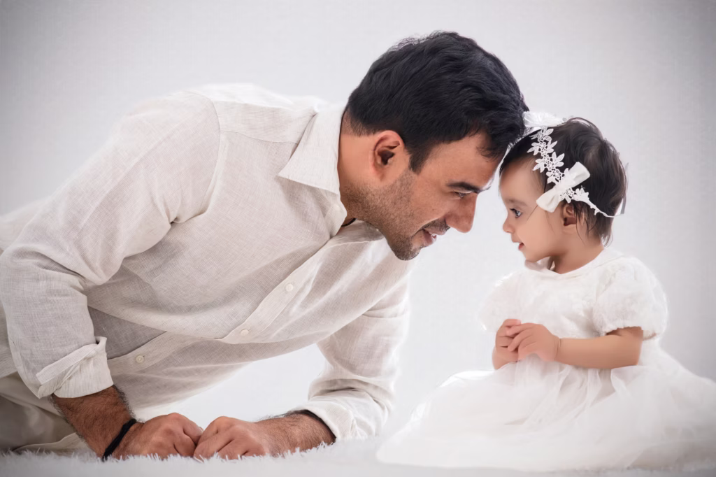 Tender indoor portrait of father and young daughter on white shag rug, father in light-colored linen shirt lying down touching forehead to daughter's, daughter in white lace dress with floral headband, calm smile - newborn photographer london father daughter forehead