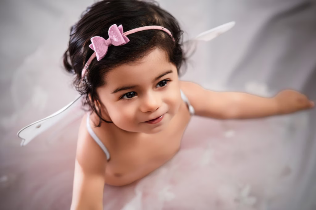 newborn photography east london High-angle portrait of toddler wearing pink bow headband and white fairy wings looking up with slight smile against softly blurred white fabric background